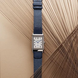 Vertical product shot of a rectangular luxury watch with a blue strap set against abstract radiating threads