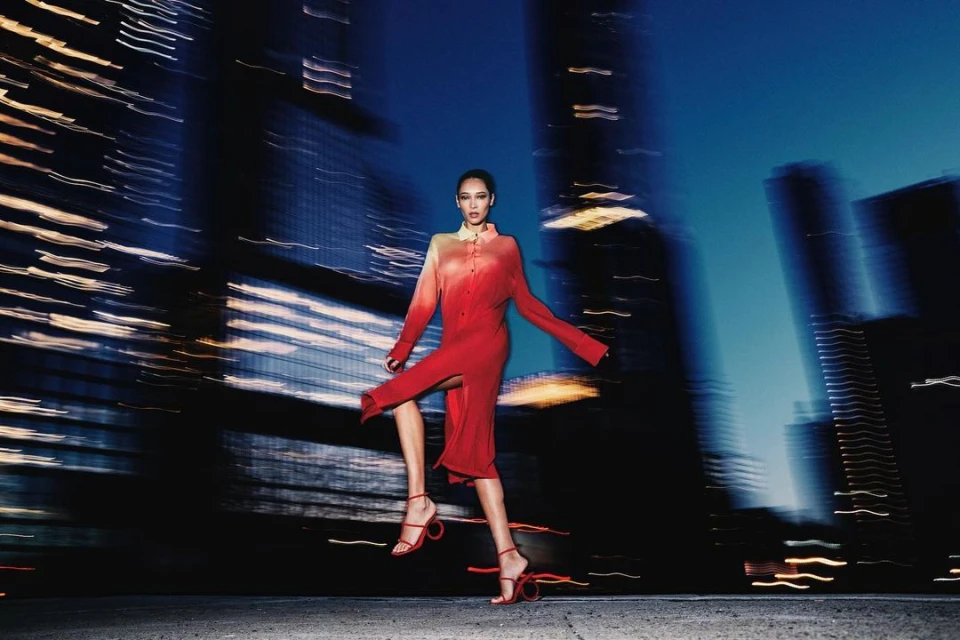 A woman in a red dress and heels poses at night with blurred city lights in the background, creating a dynamic, fashion-forward shot.