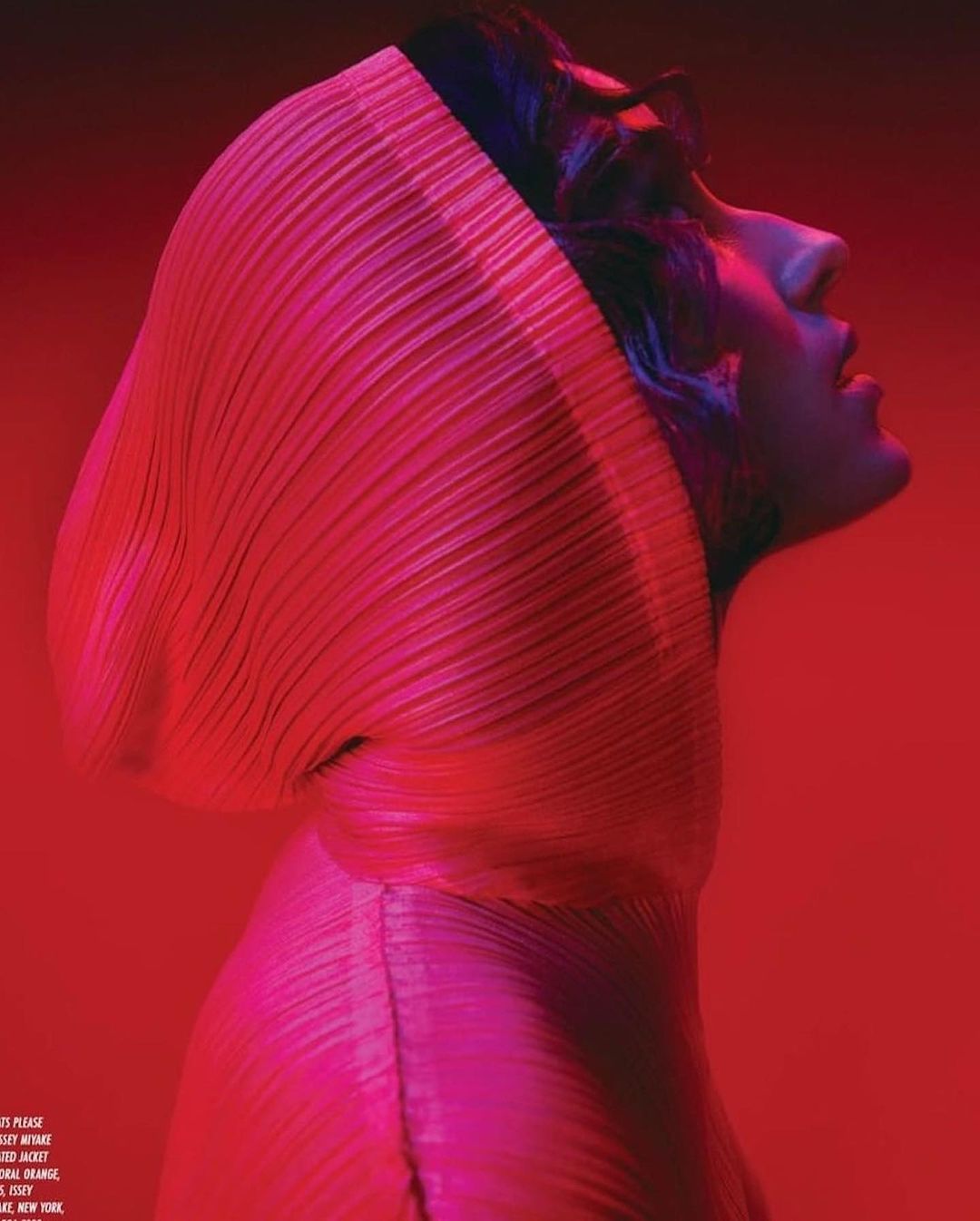 Profile of a person wearing a pleated hooded garment, posing in front of a red background, lit with pink hues, for a fashion editorial.