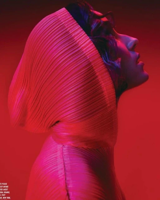 Profile of a person wearing a pleated hooded garment, posing in front of a red background, lit with pink hues, for a fashion editorial.