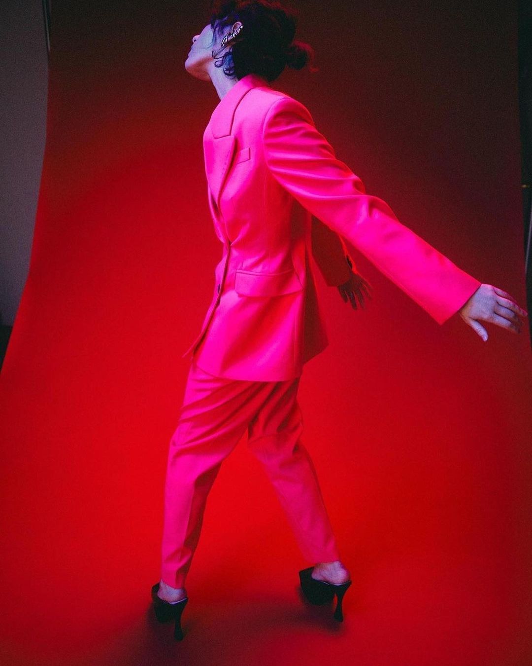 Woman poses in a bright pink suit on a matching pink background. She wears black high heels and has an accessory in her dark hair.