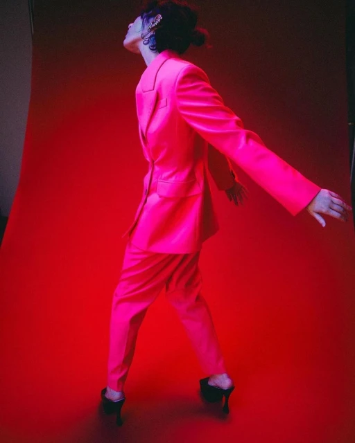 Woman poses in a bright pink suit on a matching pink background. She wears black high heels and has an accessory in her dark hair.
