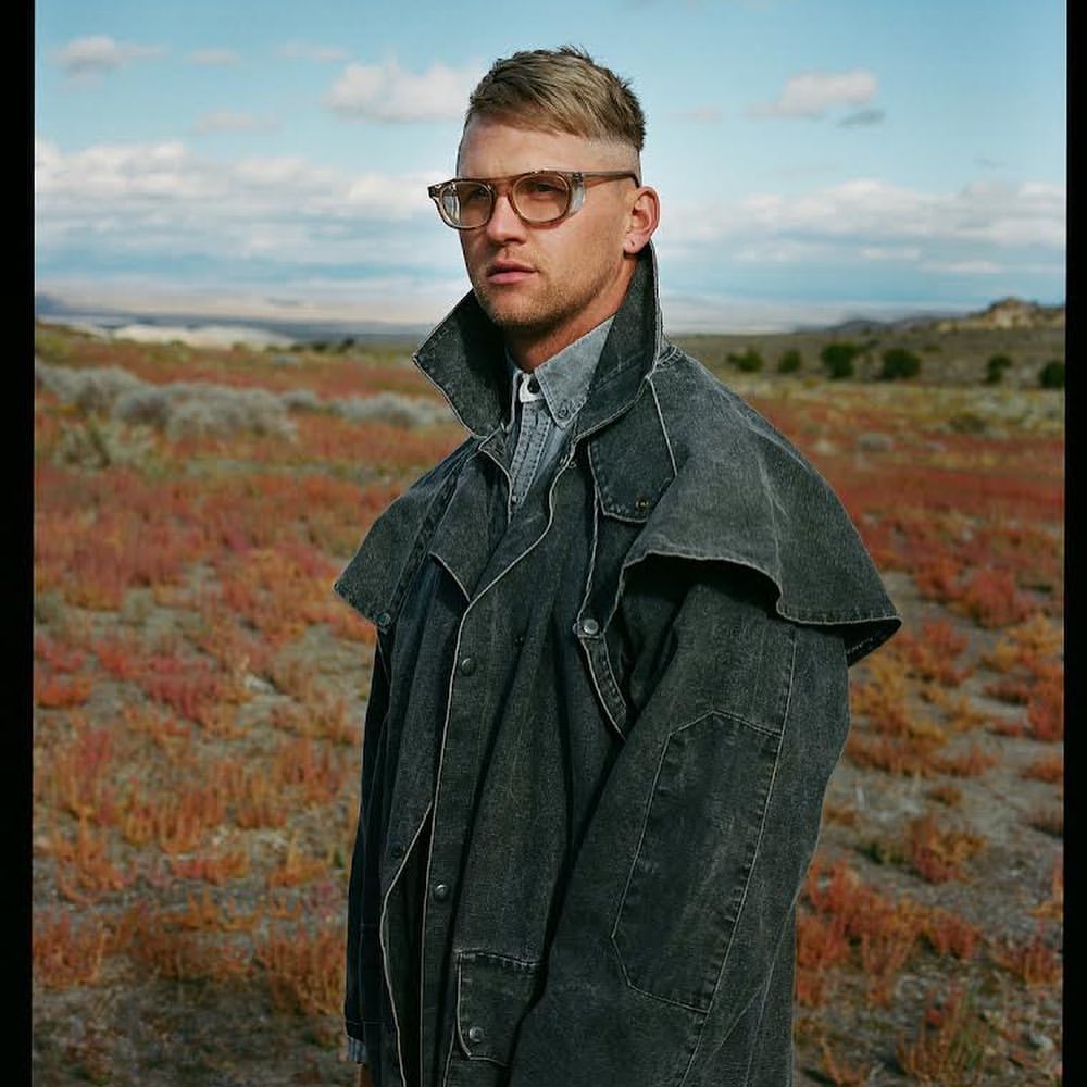 Portrait of a man in a denim duster coat and glasses, posing in a rural landscape with pink vegetation and a cloudy sky.