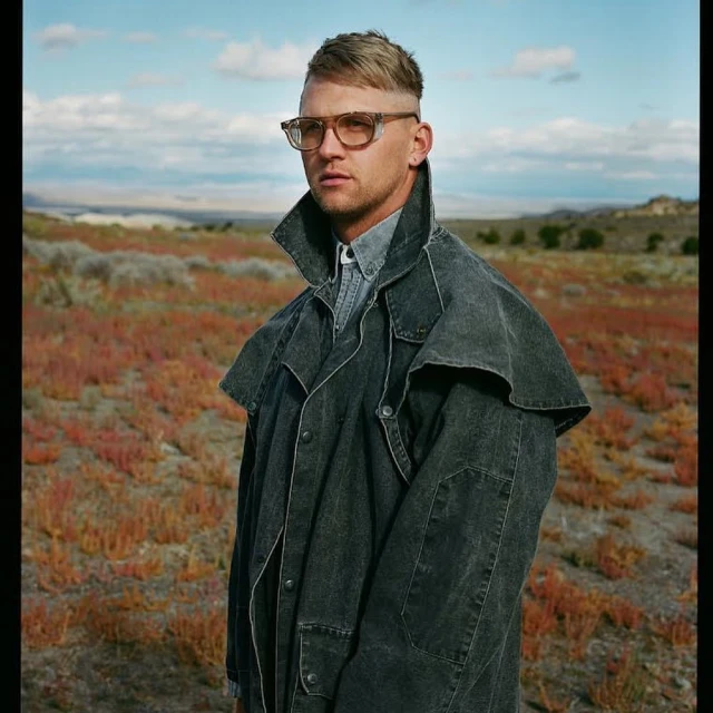 Portrait of a man in a denim duster coat and glasses, posing in a rural landscape with pink vegetation and a cloudy sky.