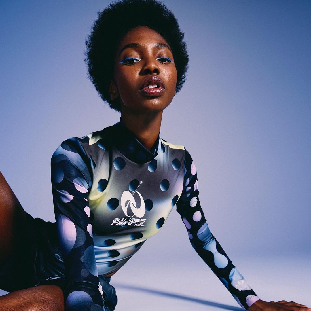 Portrait of a young Black woman with an afro wearing a patterned long-sleeve top and dark bottoms against a blue background.