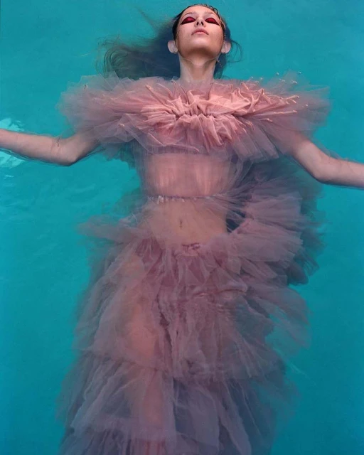 Woman in pink tulle dress floats in turquoise pool, eyes closed, arms outstretched, creating ethereal and dreamlike underwater fashion.