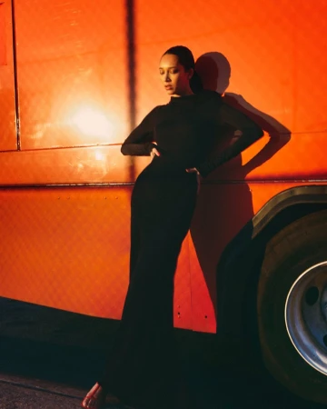 Elegant woman in a black dress poses against a vibrant orange vehicle, casting a shadow in a fashion-focused outdoor shot.