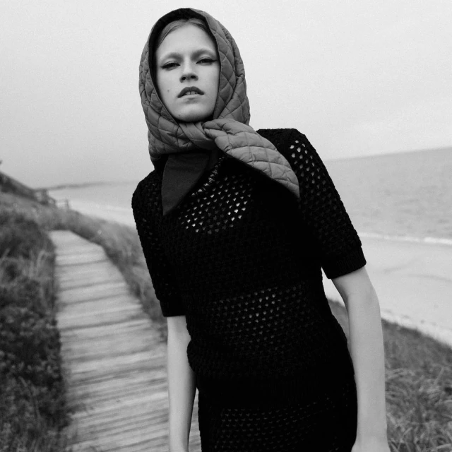 Monochrome fashion shot of a woman in a net dress and headscarf posing on a wooden path near a beach under an overcast sky.