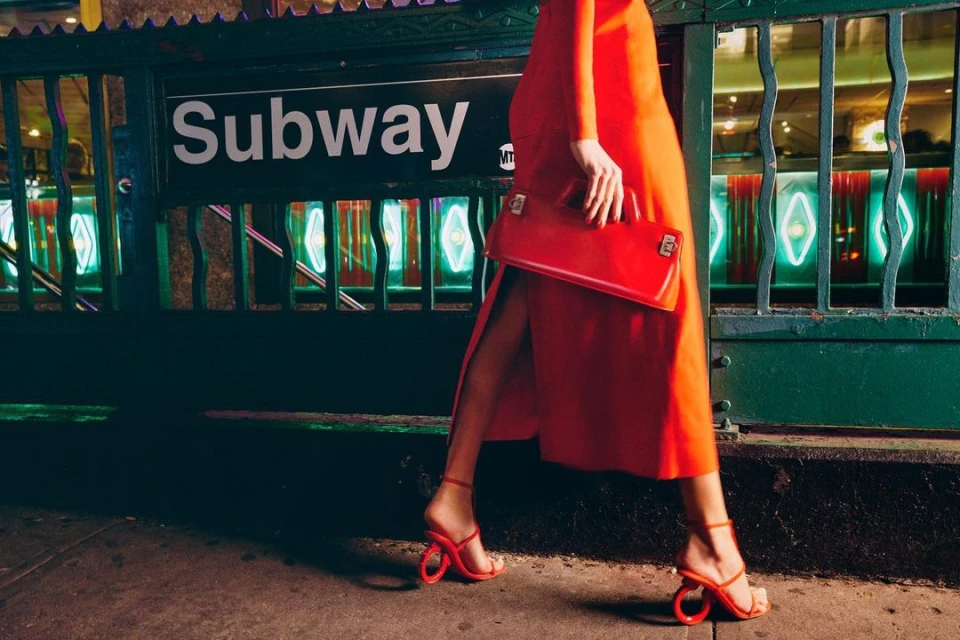Woman in a red dress and shoes walks by a subway entrance holding a red clutch. The background has subway station tiles.