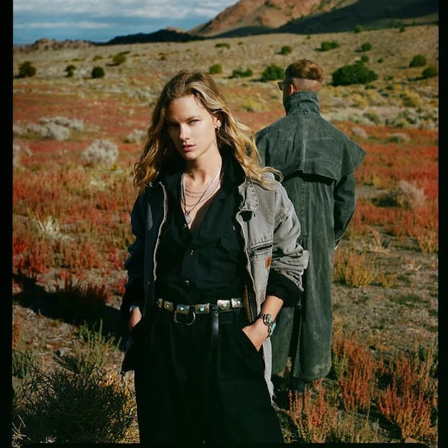 Two people in the desert wearing Western-style fashion. A woman with blonde hair stands in front of a man in a duster coat.