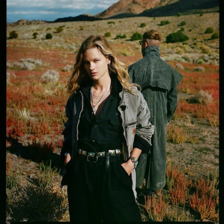 Two people in the desert wearing Western-style fashion. A woman with blonde hair stands in front of a man in a duster coat.