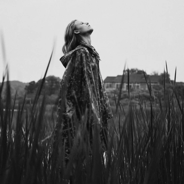 Woman in a patterned rain jacket stands in tall grass, gazing upward toward the sky with a house and trees in the background.