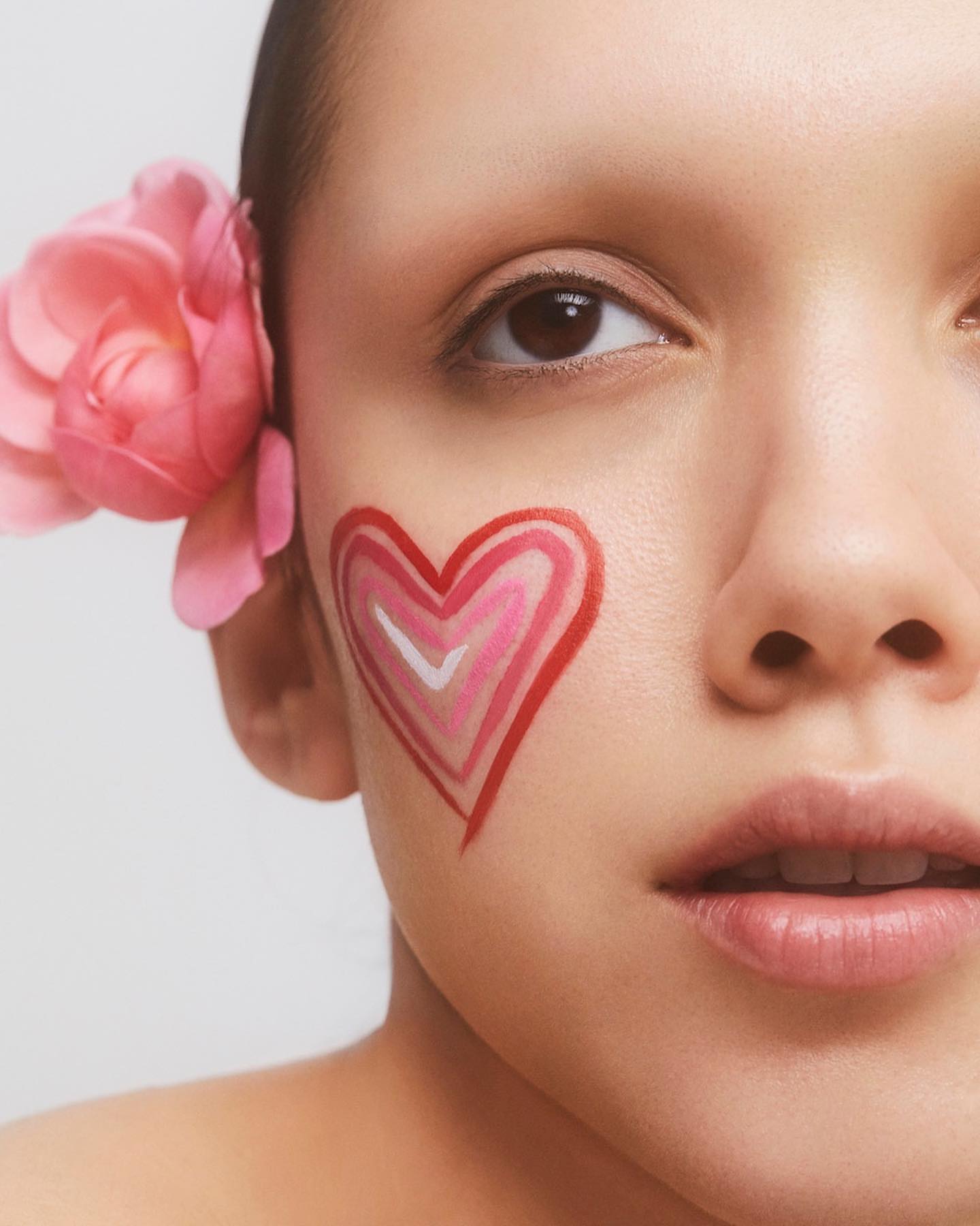 Close-up of woman's face with heart-shaped makeup and pink flower in hair against a plain white background in a beauty shot.
