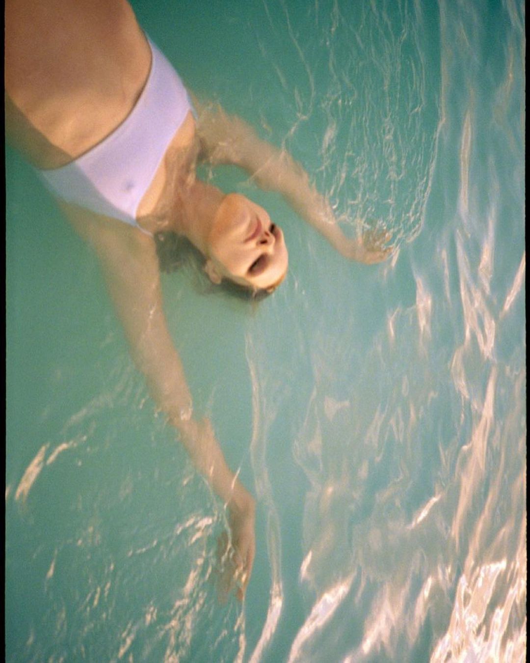 Aerial view of a woman in a light blue swimsuit floating on her back in light blue water, relaxed with her arms outstretched.
