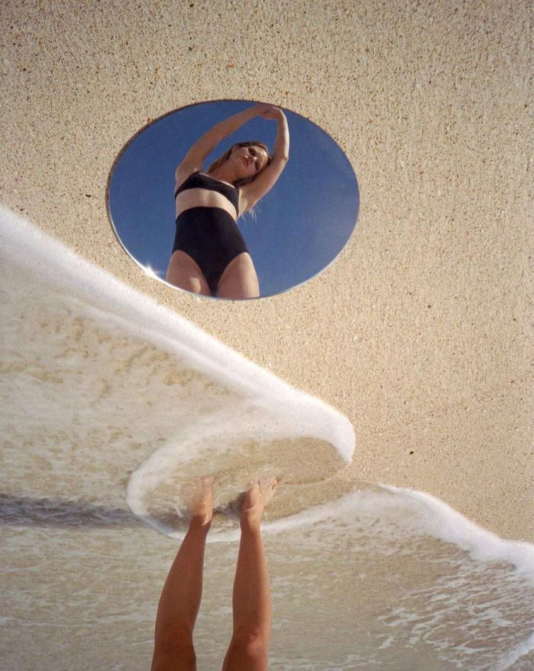 Woman in a black swimsuit reflected in a round mirror on a beach, with wave foam near her feet, creating an abstract and summery visual.