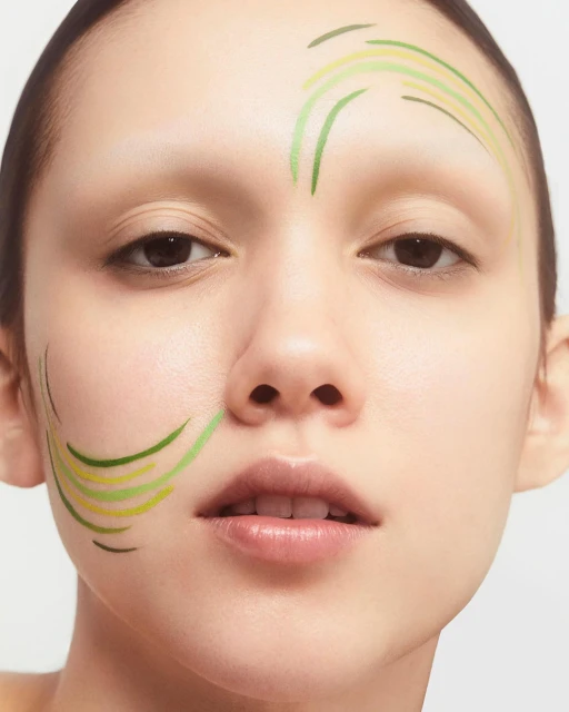 Close-up shot of a woman's face featuring green and yellow curved lines of makeup on her forehead and cheek against a white background.