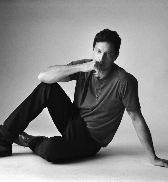 Monochrome portrait of a man with dark hair and a grey t-shirt sitting on the floor in a photo studio looking at the camera.
