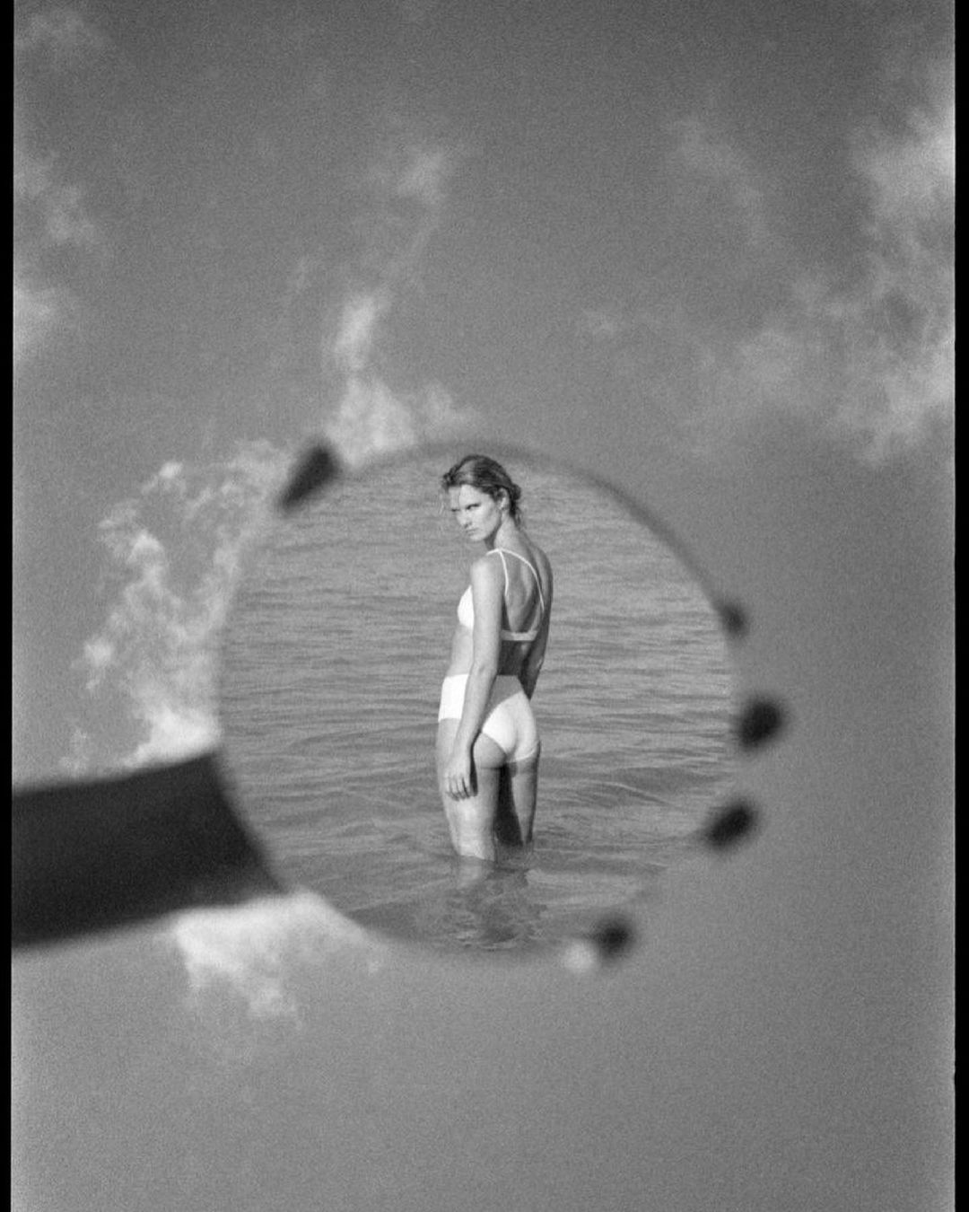 Woman in swimwear standing in water, reflected in round mirror held up by hand, with cloudy sky, black and white aesthetic.