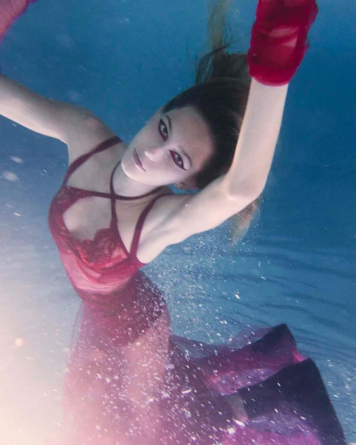 A woman swims underwater wearing a maroon dress and matching gloves, posing in a clear blue pool with visible bubbles.
