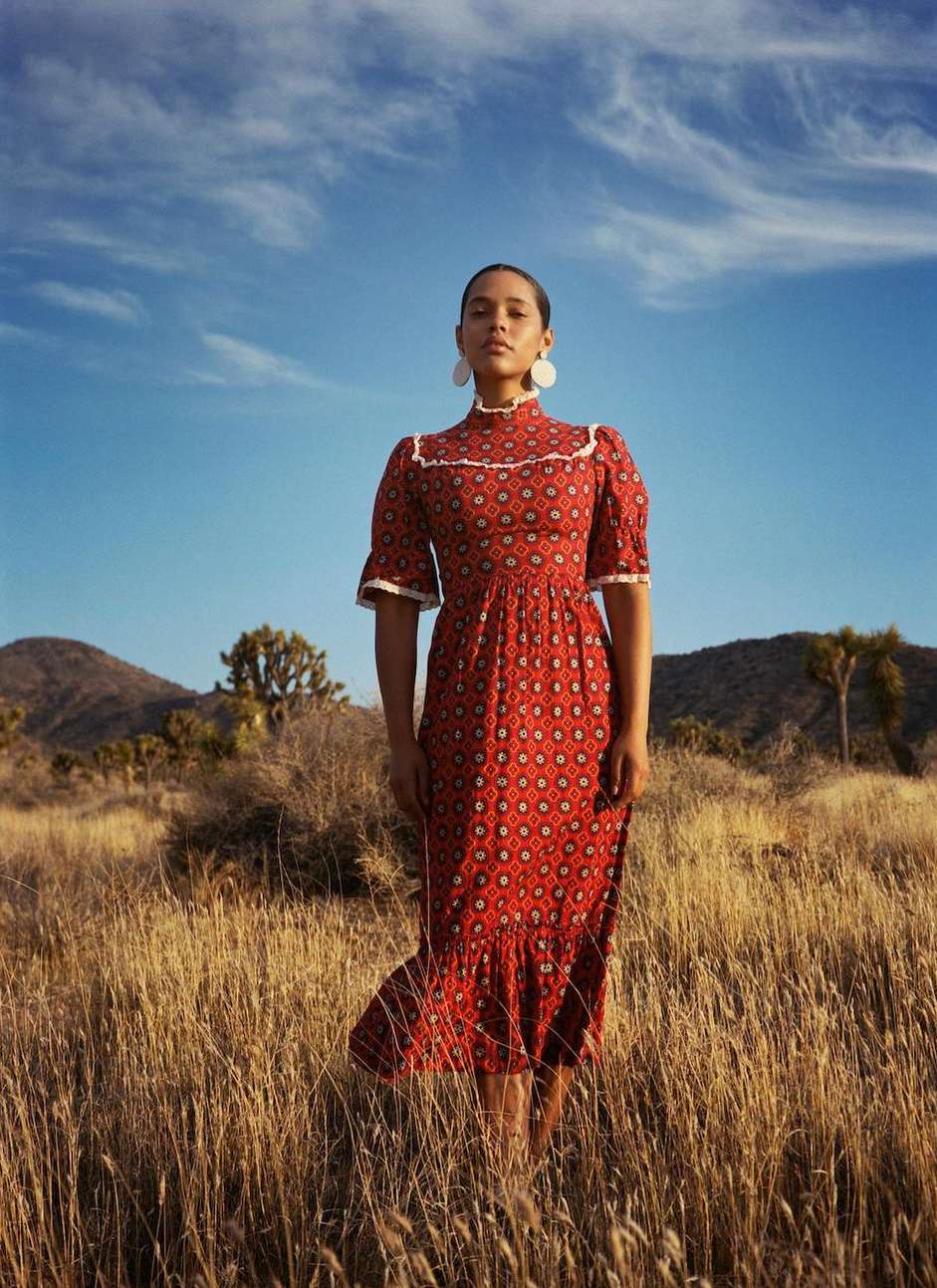 A woman in a patterned dress stands in a field of tall grass against a backdrop of mountains and a blue sky with light clouds.