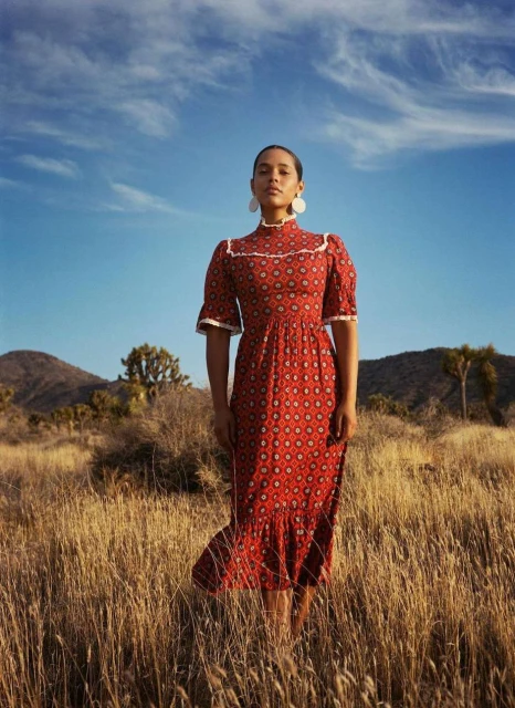 A woman in a patterned dress stands in a field of tall grass against a backdrop of mountains and a blue sky with light clouds.