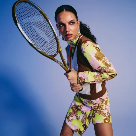 A young adult female model poses in floral print sportswear while holding a tennis racket in a studio setting with a blue background.