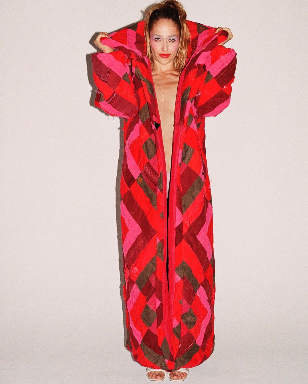 Woman in a long red and pink quilted coat stands against a white background in a studio setting, capturing a unique fashion statement.
