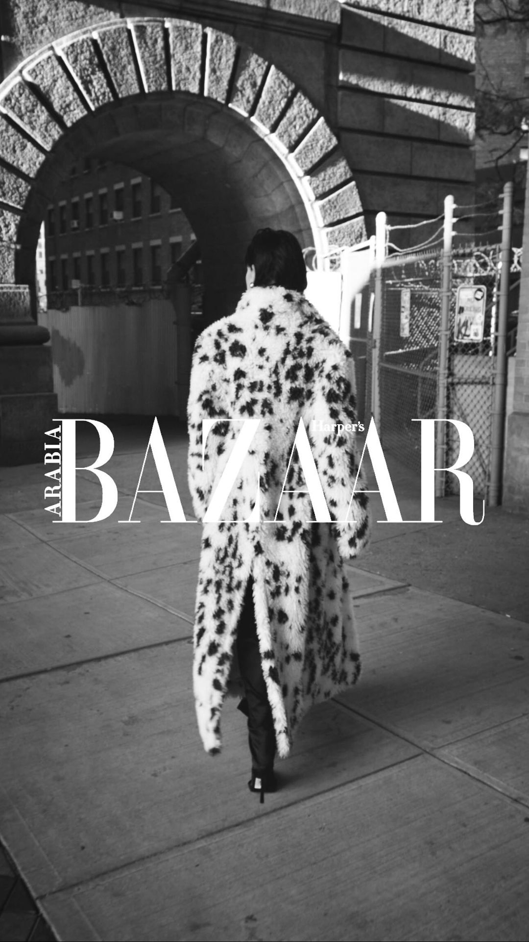 Monochrome fashion shot of a person in a spotted faux fur coat walking on a sidewalk, with an arched stone building in the background.
