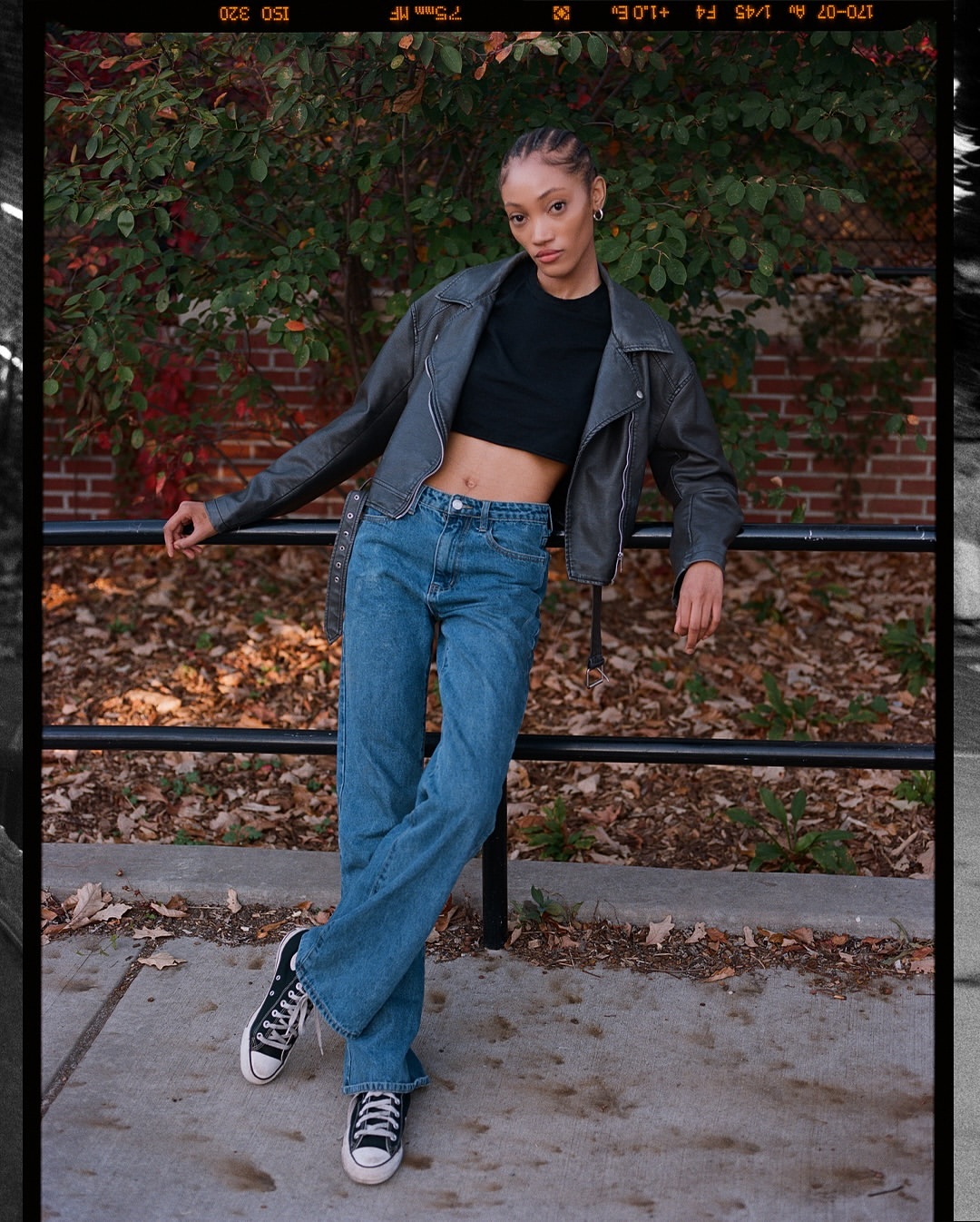 A young adult woman poses outdoors wearing a gray leather jacket, black crop top, blue jeans, and black sneakers in natural light.