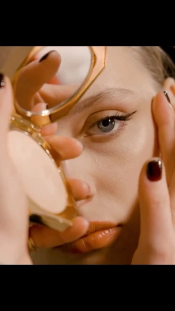 Close-up shot of a fair-skinned adult woman's face as she holds a compact mirror, showcasing her makeup and red-painted fingernails.