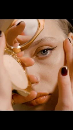 Close-up shot of a fair-skinned adult woman's face as she holds a compact mirror, showcasing her makeup and red-painted fingernails.