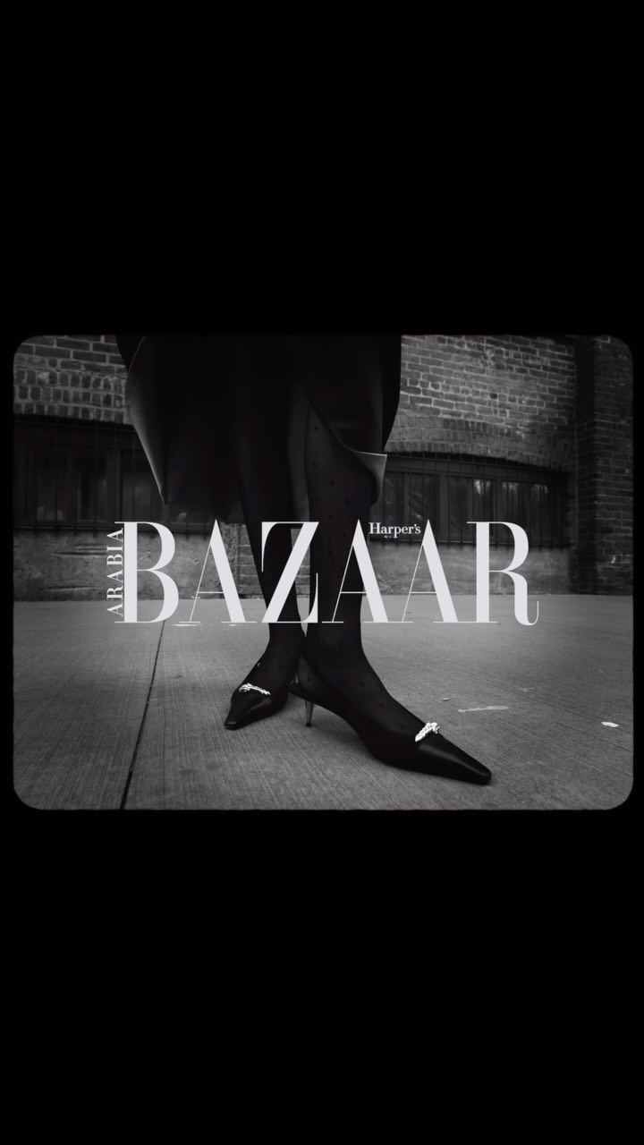 A black-and-white fashion shot featuring a woman's legs and feet in black pointed-toe heels against a brick building and concrete sidewalk.