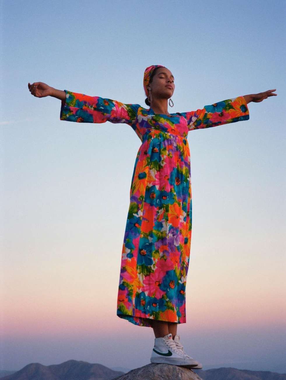A woman in a colorful floral dress stands on a rock with arms outstretched against a blue sky and distant mountains at sunset.