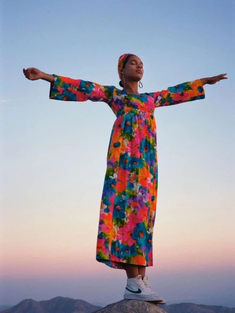 A woman in a colorful floral dress stands on a rock with arms outstretched against a blue sky and distant mountains at sunset.