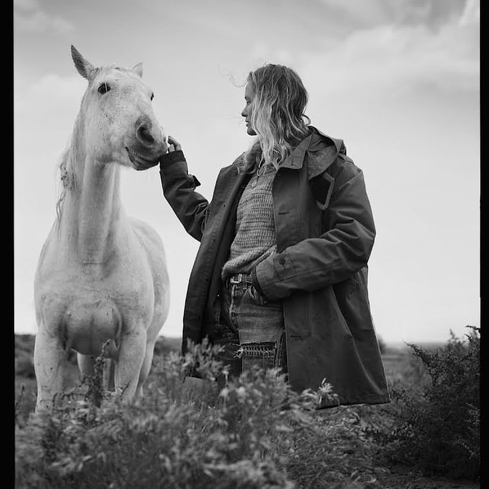 Black-and-white photo of a woman in a coat gently touching the face of a white horse in an outdoor, grassy field under a cloudy sky.