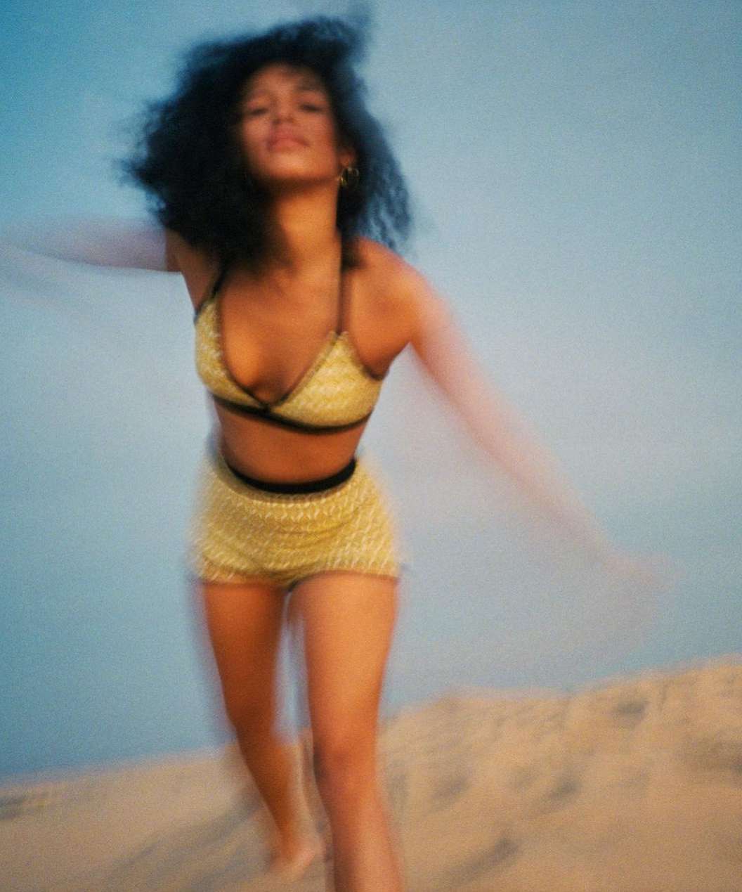 Woman in yellow bikini and shorts poses with arms outstretched on a sandy beach against a light blue sky. Motion blur effect.