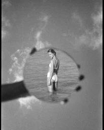 Monochrome image of a woman in a white bikini standing in shallow water, viewed through a circular handheld mirror.