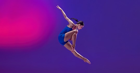 Solo female dancer in blue leotard mid-air against a split pink and purple gradient background performing a dance move.