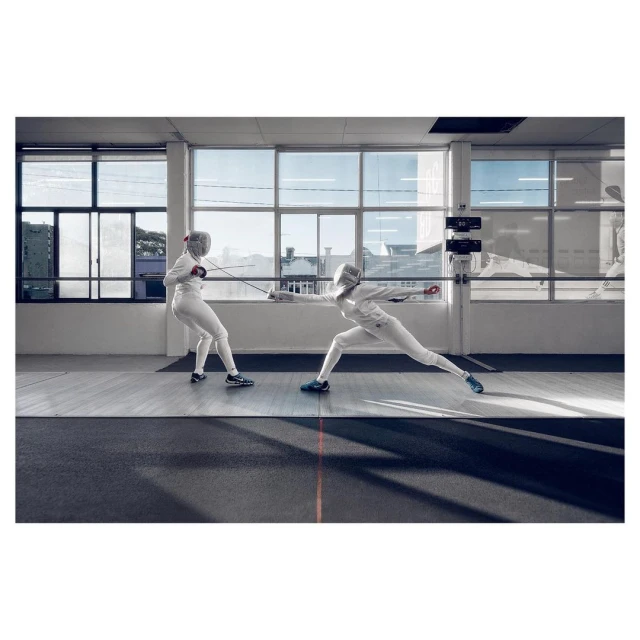 Two fencers in white uniforms compete in a brightly lit indoor arena, lunging with foils, wearing protective masks, and displaying athletic skill.