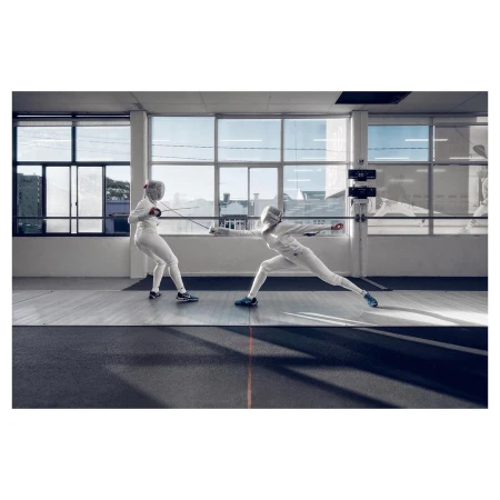 Two fencers in white uniforms compete in a brightly lit indoor arena, lunging with foils, wearing protective masks, and displaying athletic skill.