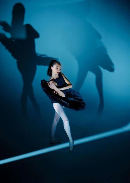 A ballerina in a black tutu poses en pointe in a dance studio with dramatic shadows, creating an elegant and artistic dance performance.