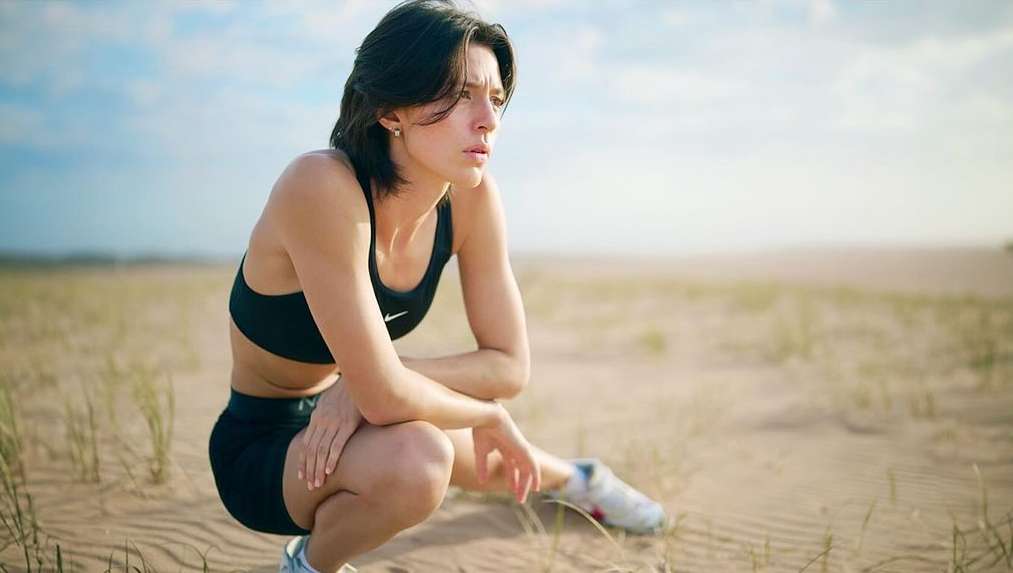 Female athlete in sports bra and shorts crouches on sandy ground with a distant blue sky, looking to the side in an outdoor environment.