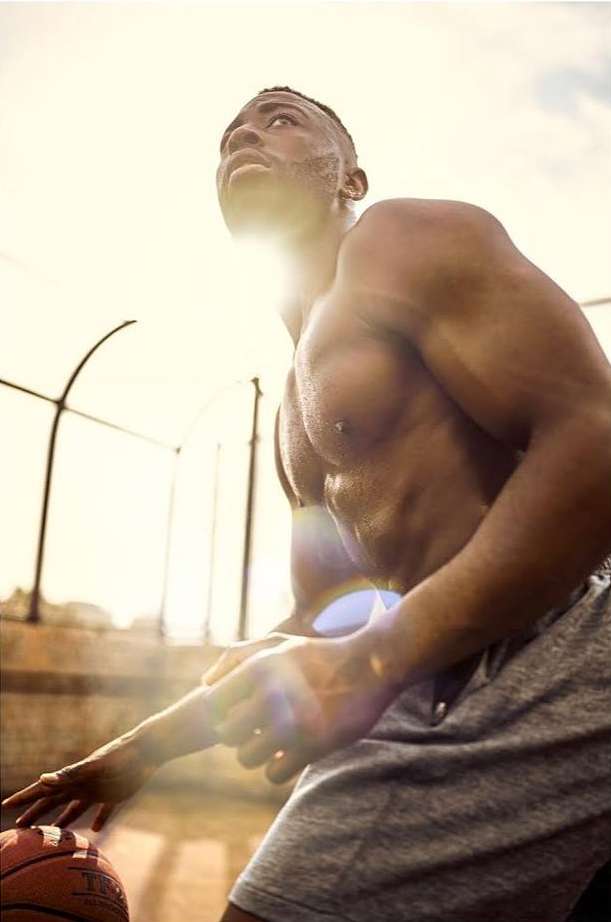 Athletic African-American man dribbling a basketball outdoors in bright sunlight. Muscular build, gray shorts, and focused gaze.