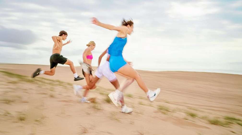 Group of four people running on a sand dune under a cloudy sky, with motion blur enhancing the dynamic sense of movement in an outdoor setting.