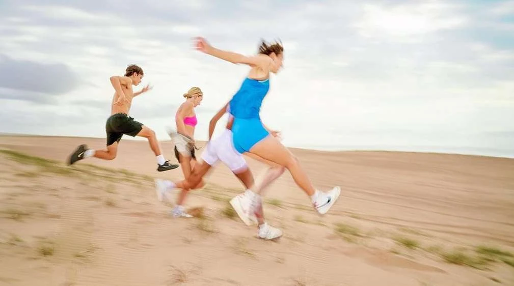 Group of four people running on a sand dune under a cloudy sky, with motion blur enhancing the dynamic sense of movement in an outdoor setting.