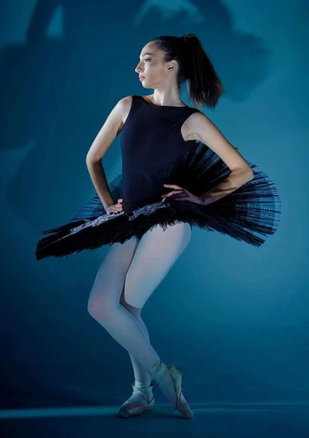 Full shot of a ballerina in a black tutu and ballet slippers posing in a dance studio with a blue background in a studio shot.