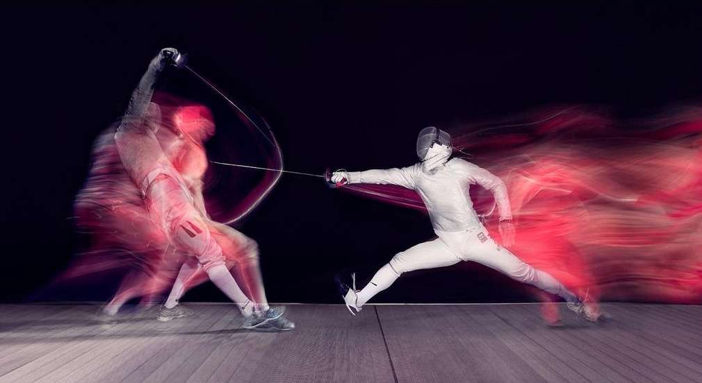 Two fencers in white uniforms engaged in a bout, motion blur conveys fast-paced action, set against a dark background.