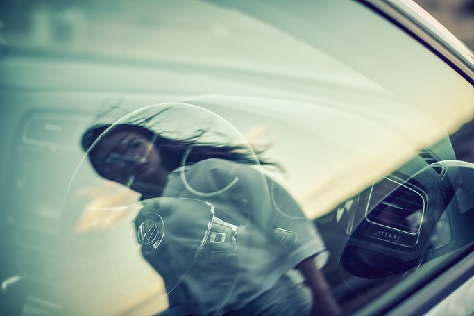View through car windshield of woman with long dark hair in motion behind the wheel; Volkswagen emblem visible on steering wheel.