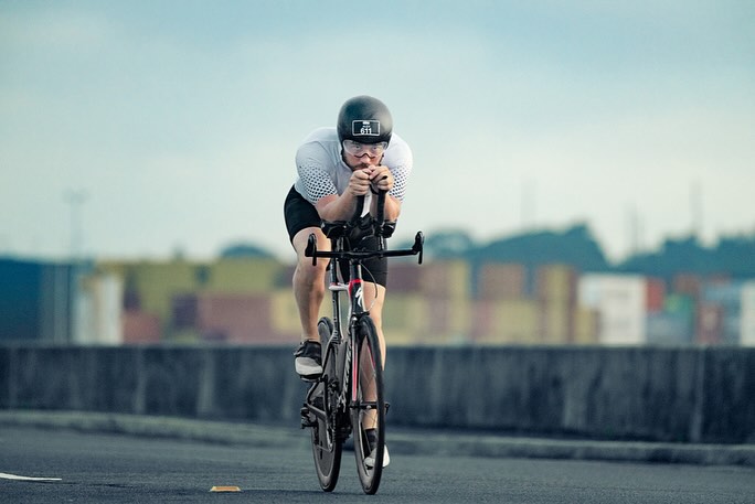A cyclist in a helmet and sporting gear rides a racing bike on an asphalt road. Cargo containers are visible in the background.