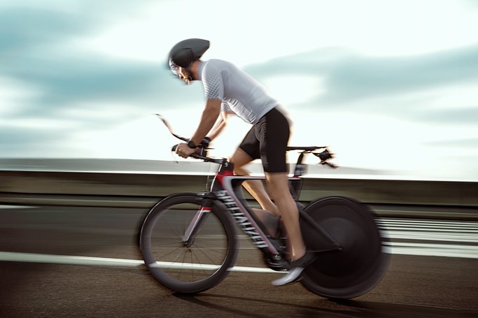 A cyclist races on a Specialized road bike along a highway, showcasing speed with motion blur against a cloudy sky and concrete barriers.